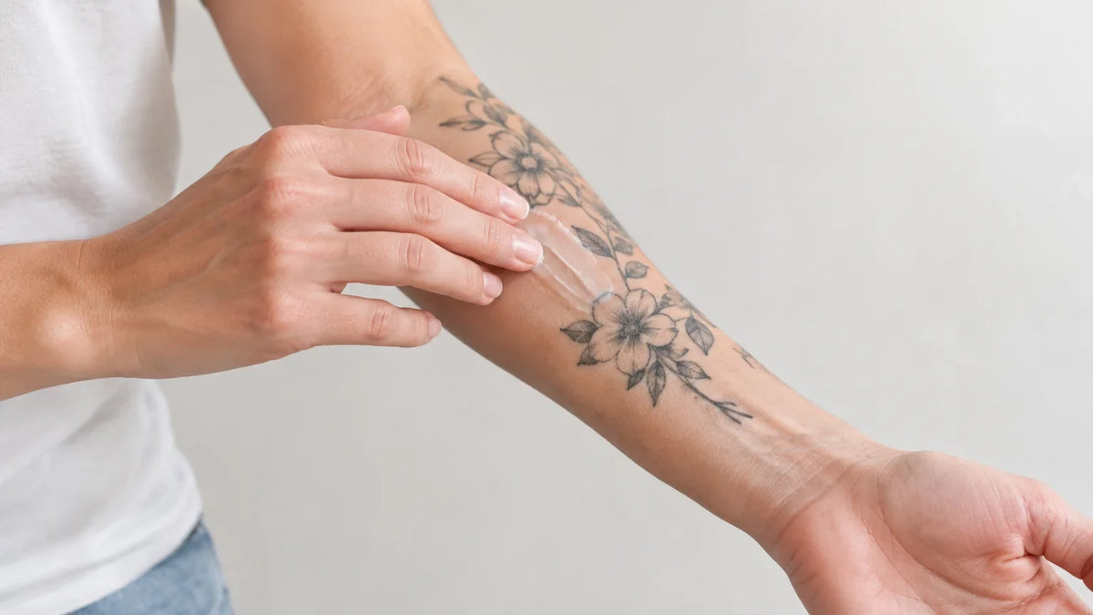 Une personne prend soin d’un tatouage sur son avant-bras avec un baume, dans une salle de bain lumineuse. Image
